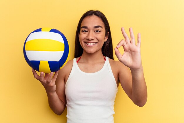 Giovane donna di razza mista che gioca a pallavolo sulla spiaggia isolata su sfondo giallo allegro e fiducioso che mostra gesto ok.