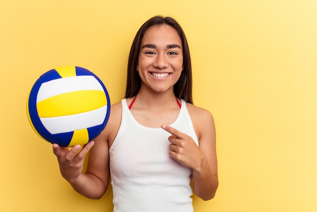 Giovane donna di razza mista che gioca a pallavolo sulla spiaggia isolata su sfondo giallo sorridendo e indicando da parte, mostrando qualcosa in uno spazio vuoto.