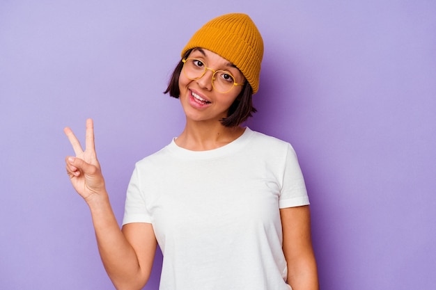 Giovane donna di razza mista che indossa un berretto di lana isolato su sfondo viola gioiosa e spensierata che mostra un simbolo di pace con le dita.