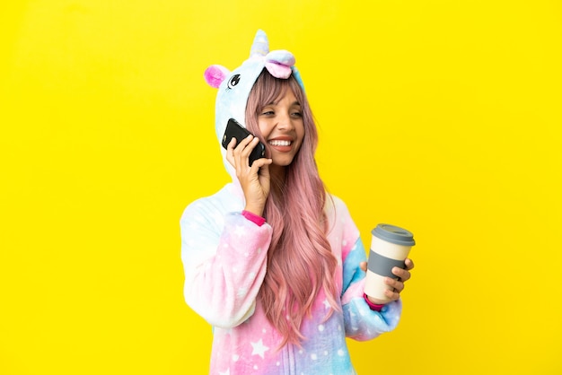 Giovane donna di razza mista che indossa un pigiama unicorno isolato su sfondo bianco che tiene il caffè da portare via e un cellulare