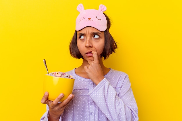 Giovane donna di razza mista che mangia cereali che indossa un pijama isolato sul muro giallo rilassato pensando a qualcosa guardando uno spazio di copia.