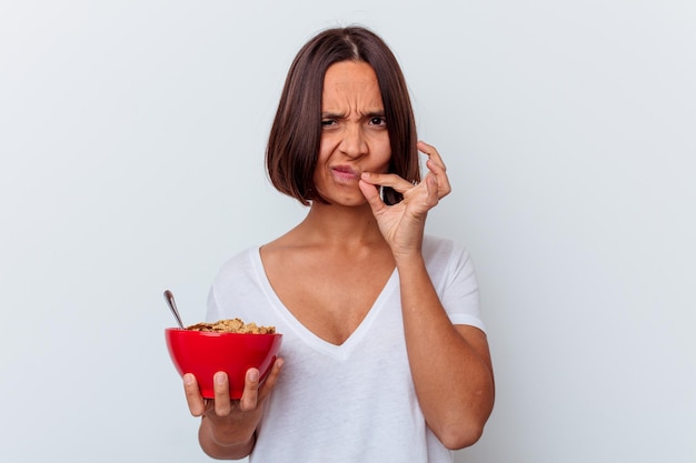 Giovane donna di razza mista che mangia cereali isolato sul muro bianco con le dita sulle labbra mantenendo un segreto.