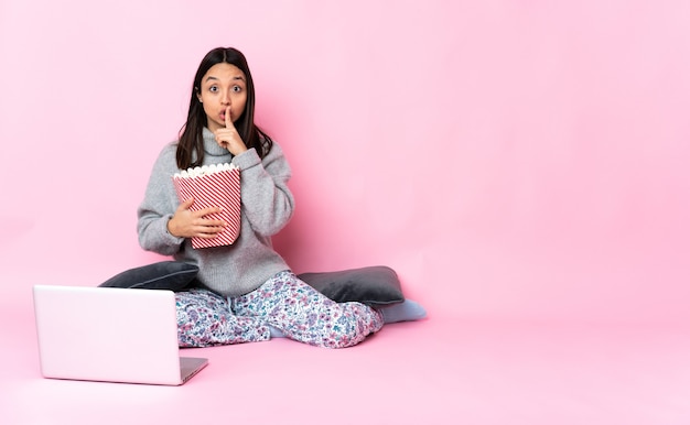 Giovane donna di razza mista che mangia popcorn mentre guarda un film sul laptop che punta di lato e fa il gesto del silenzio