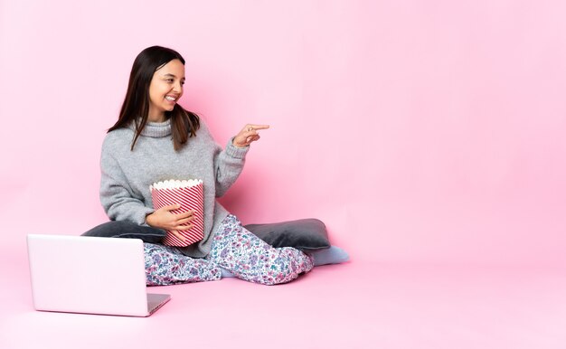 Giovane donna di razza mista che mangia popcorn mentre guarda un film sul laptop che punta il dito di lato e presenta un prodotto