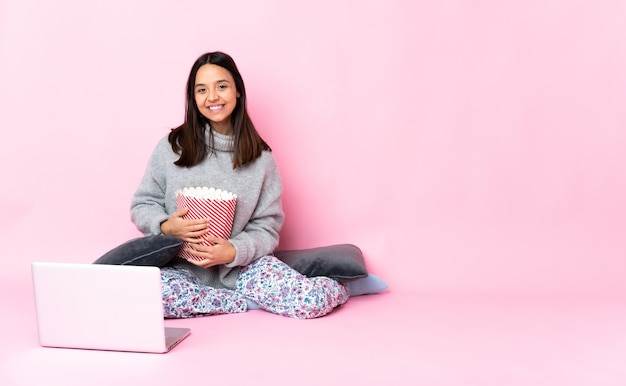 Giovane donna di razza mista che mangia popcorn mentre guarda un film sul laptop mantenendo le braccia incrociate in posizione frontale