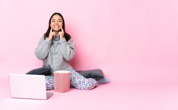 Giovane donna di razza mista che mangia popcorn mentre guarda un film sul laptop sorridendo con un'espressione felice e piacevole