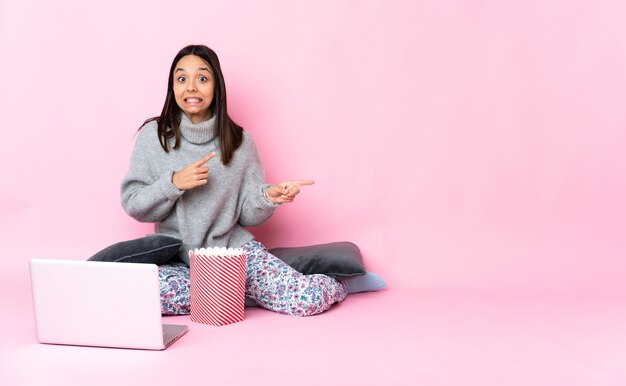 Giovane donna di razza mista che mangia popcorn mentre guarda un film sul laptop spaventata e indicando il lato