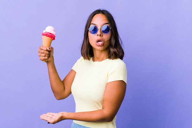 Giovane donna di razza mista che mangia un gelato scioccata e stupita tenendo uno spazio di copia tra le mani.