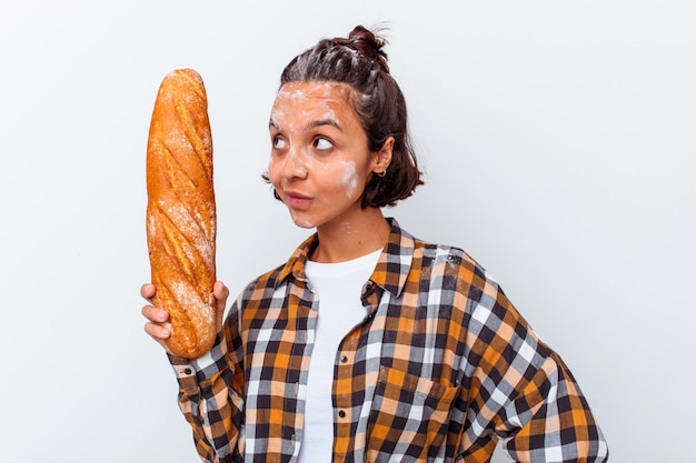 Giovane donna di razza mista che produce pane isolato sul muro bianco guarda da parte sorridente, allegro e piacevole.