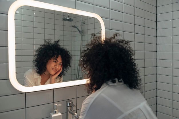Giovane donna di razza mista che si guarda allo specchio con amore in bagno Concetto di amore di sé