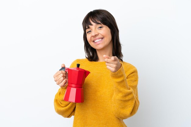 Giovane donna di razza mista che tiene caffettiera isolata su sfondo bianco che punta davanti con espressione felice