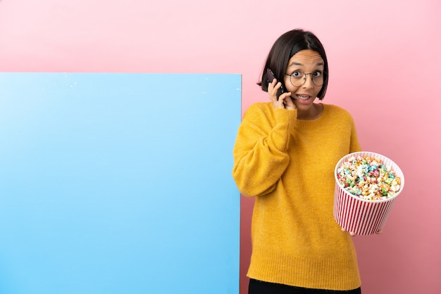 Giovane donna di razza mista che tiene i popcorn con un grande striscione su sfondo isolato mantenendo una conversazione con il telefono cellulare con qualcuno