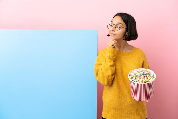 Giovane donna di razza mista che tiene i popcorn con un grande striscione su sfondo isolato pensando un'idea mentre guarda in alto