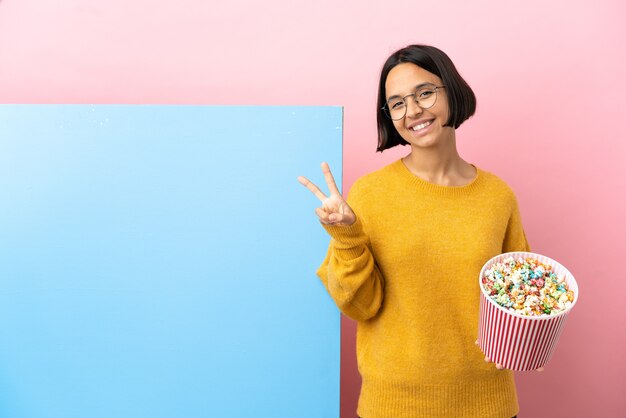 Giovane donna di razza mista che tiene i popcorn con un grande striscione su sfondo isolato sorridendo e mostrando il segno della vittoria