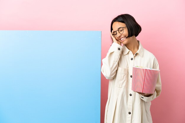 Giovane donna di razza mista che tiene i popcorn con un grande striscione su sfondo isolato sorridente molto