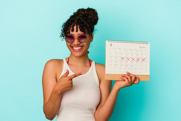 Giovane donna di razza mista che tiene il calendario isolato su sfondo blu sorridente e puntato da parte, mostrando qualcosa in uno spazio vuoto.