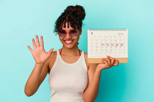 Giovane donna di razza mista che tiene il calendario isolato sul muro blu sorridente allegro che mostra il numero cinque con le dita.