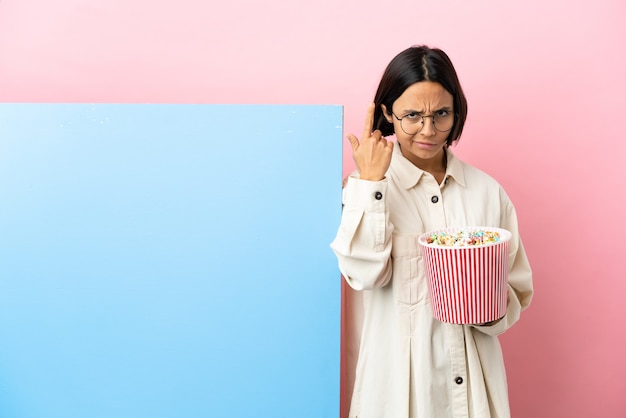 Giovane donna di razza mista che tiene in mano i popcorn con un grande striscione su sfondo isolato che fa il gesto della follia mettendo il dito sulla testa