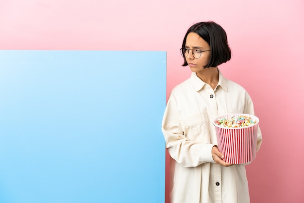 Giovane donna di razza mista che tiene in mano i popcorn con un grande striscione su sfondo isolato che ha dubbi mentre guarda di lato