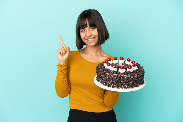 Giovane donna di razza mista che tiene in mano una torta di compleanno che mostra e alza un dito in segno del meglio