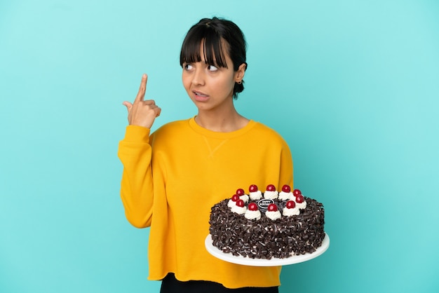 Giovane donna di razza mista che tiene la torta di compleanno facendo il gesto della follia mettendo il dito sulla testa
