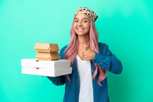 Giovane donna di razza mista che tiene pizze e hamburger isolati su sfondo verde dando un gesto di pollice in alto