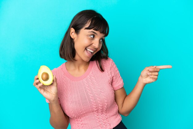 Giovane donna di razza mista che tiene un avocado isolato su sfondo blu che punta il dito di lato e presenta un prodotto