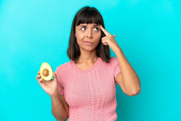 Giovane donna di razza mista che tiene un avocado isolato su sfondo blu con dubbi e pensare