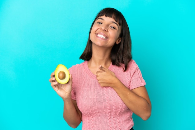 Giovane donna di razza mista che tiene un avocado isolato su sfondo blu dando un gesto di pollice in alto