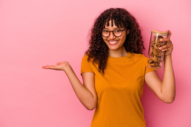 Giovane donna di razza mista che tiene un barattolo di biscotti isolato sul muro rosa che mostra uno spazio di copia su un palmo e che tiene un'altra mano sulla vita.