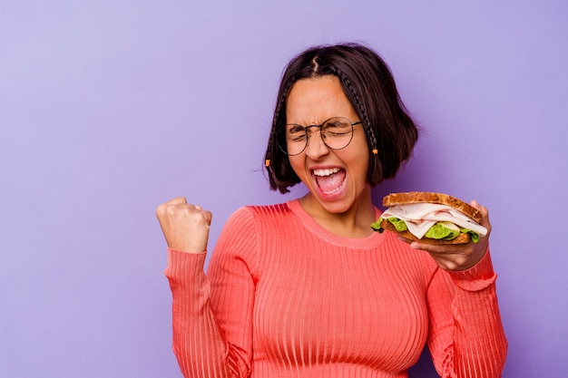 Giovane donna di razza mista che tiene un panino isolato su sfondo viola alzando il pugno dopo una vittoria, concetto di vincitore.