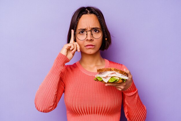 Giovane donna di razza mista che tiene un panino isolato su sfondo viola che punta il tempio con il dito, pensando, concentrato su un compito.