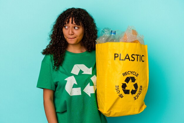 Giovane donna di razza mista che tiene un sacchetto di plastica riciclato isolato su sfondo blu confuso, si sente dubbioso e insicuro.
