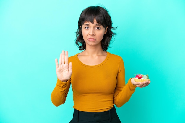 Giovane donna di razza mista che tiene un tortino isolato su sfondo blu facendo un gesto di arresto