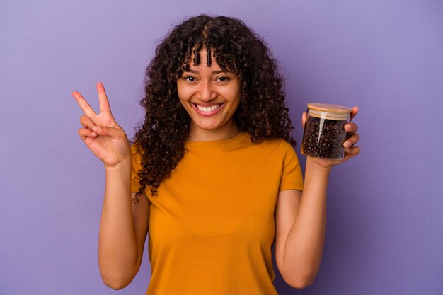 Giovane donna di razza mista che tiene una bottiglia di chicchi di caffè isolata su sfondo viola che mostra il numero due con le dita.