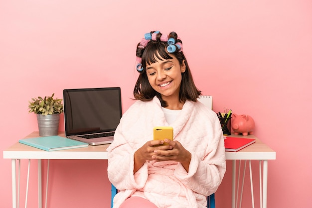 Giovane donna di razza mista con bigodini che tengono il telefono cellulare isolato su sfondo rosa