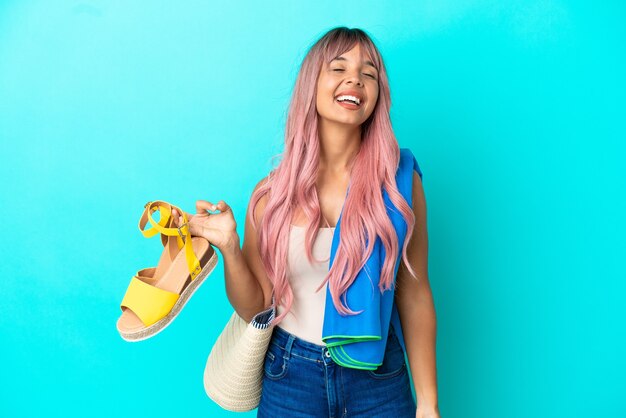 Giovane donna di razza mista con capelli rosa che tiene sandali estivi isolati su sfondo blu ridendo