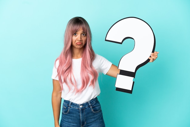 Giovane donna di razza mista con capelli rosa isolata su sfondo blu con in mano un'icona a punto interrogativo e con espressione triste
