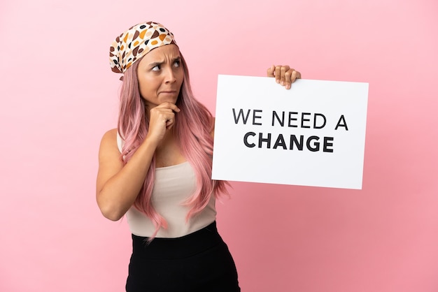 Giovane donna di razza mista con capelli rosa isolata su sfondo rosa con in mano un cartello con testo Abbiamo bisogno di un cambiamento e di pensare