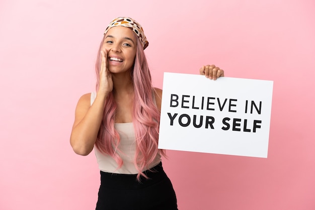 Giovane donna di razza mista con capelli rosa isolati su sfondo rosa con in mano un cartello con testo Credi in te stesso e urla