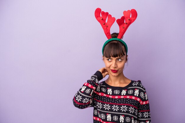 Giovane donna di razza mista con cappello da renna che celebra il Natale isolato su sfondo viola toccando la parte posteriore della testa, pensando e facendo una scelta.