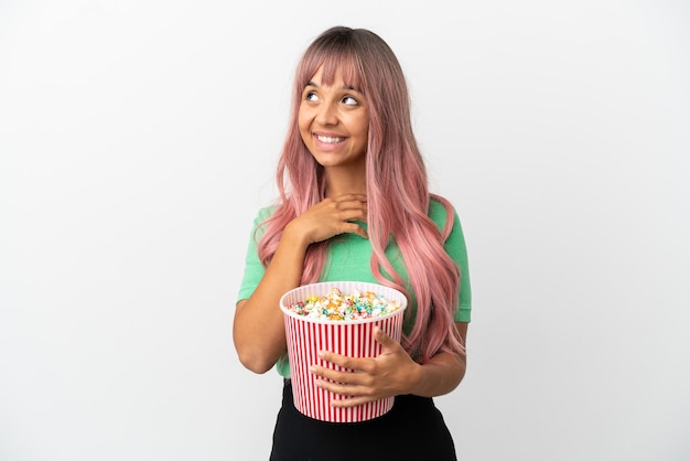 Giovane donna di razza mista con i capelli rosa che mangia popcorn isolato su sfondo bianco alzando lo sguardo mentre sorride