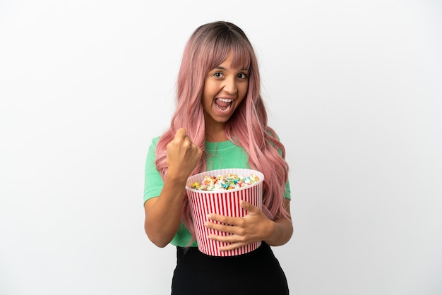 Giovane donna di razza mista con i capelli rosa che mangia popcorn isolato su sfondo bianco che celebra una vittoria