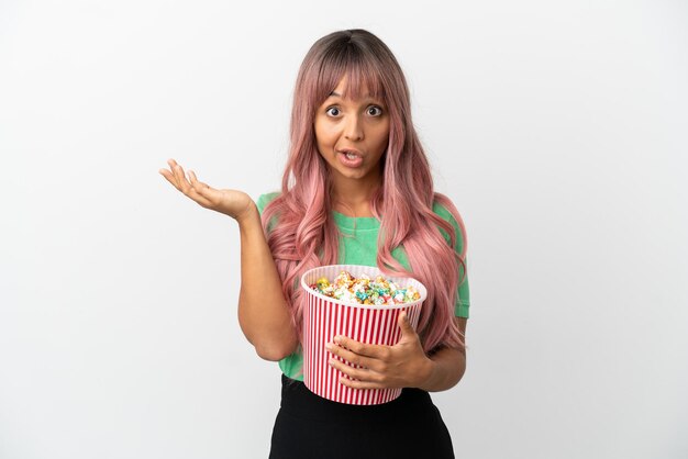 Giovane donna di razza mista con i capelli rosa che mangia popcorn isolato su sfondo bianco con espressione facciale scioccata