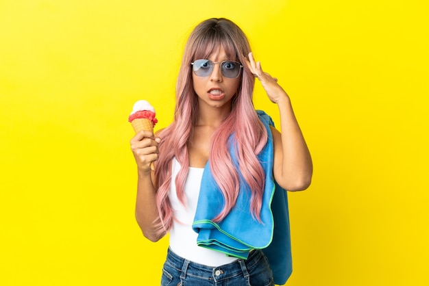 Giovane donna di razza mista con i capelli rosa che tiene il gelato isolato su sfondo giallo con espressione a sorpresa