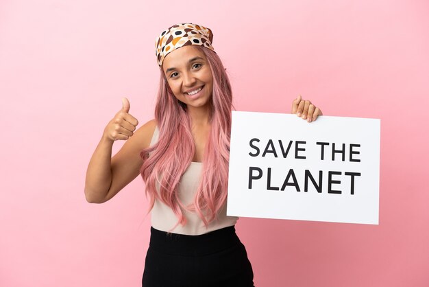 Giovane donna di razza mista con i capelli rosa isolata su sfondo rosa che tiene un cartello con il testo Salva il pianeta con il pollice in su