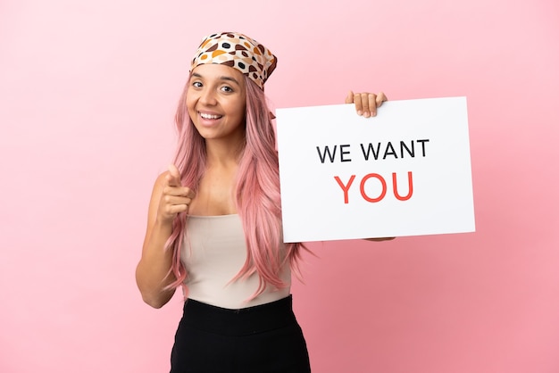 Giovane donna di razza mista con i capelli rosa isolata su sfondo rosa tenendo We Want You board e puntando verso la parte anteriore