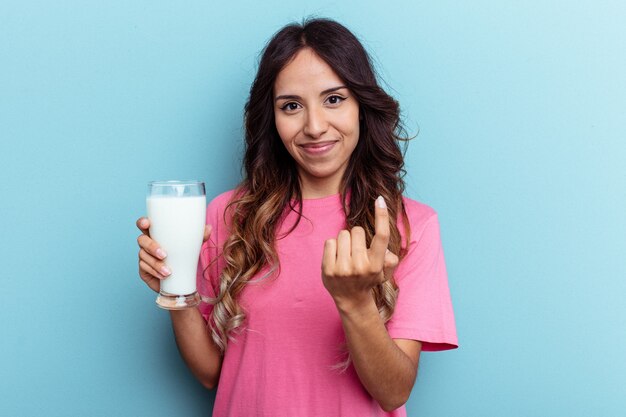 Giovane donna di razza mista con in mano un bicchiere di latte isolato su sfondo blu che punta con il dito verso di te come se invitasse ad avvicinarsi.