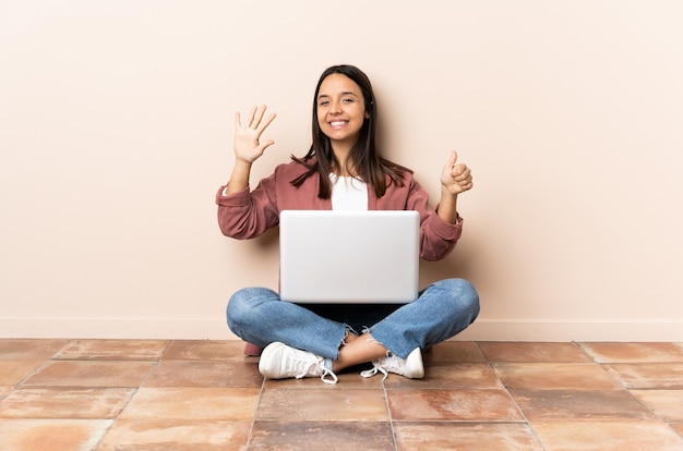 Giovane donna di razza mista con un laptop seduto sul pavimento contando sei con le dita