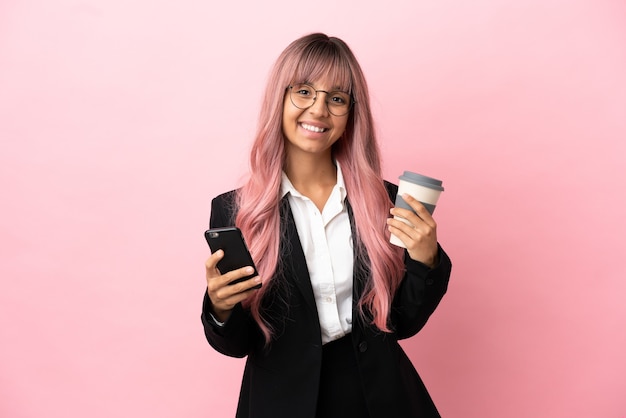 Giovane donna di razza mista d'affari con i capelli rosa isolata su sfondo rosa che tiene il caffè da portare via e un cellulare